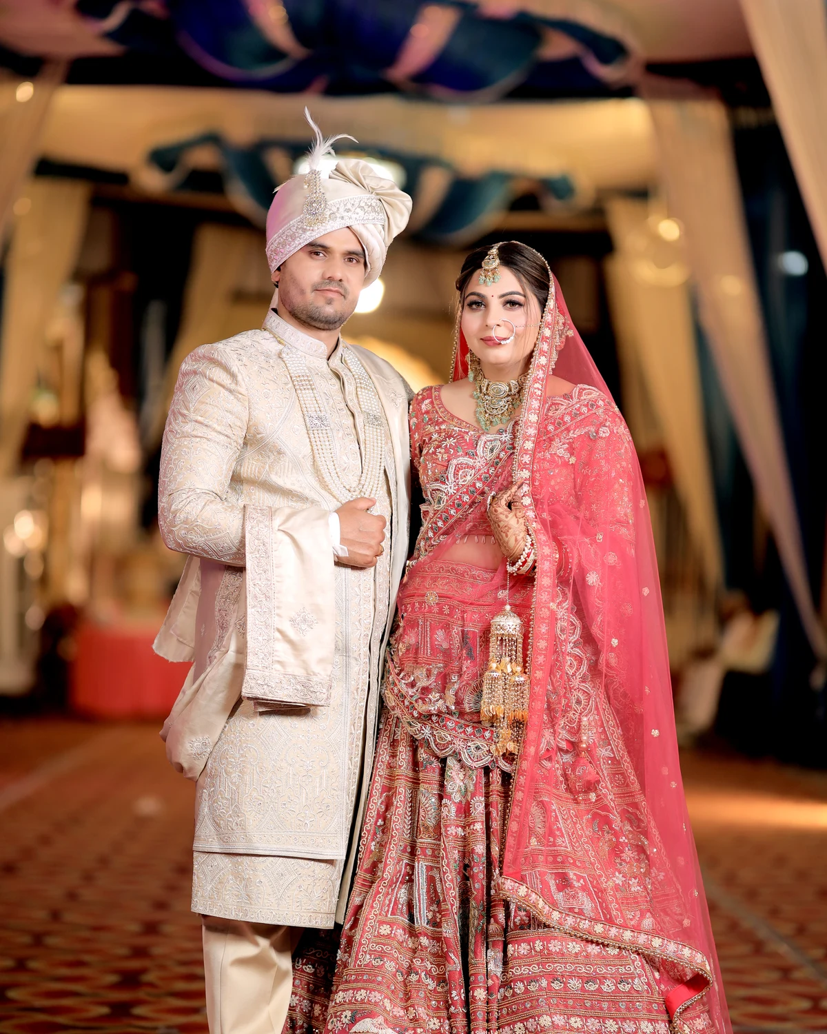 Groom in an ivory sherwani beside his bride in a grand hall