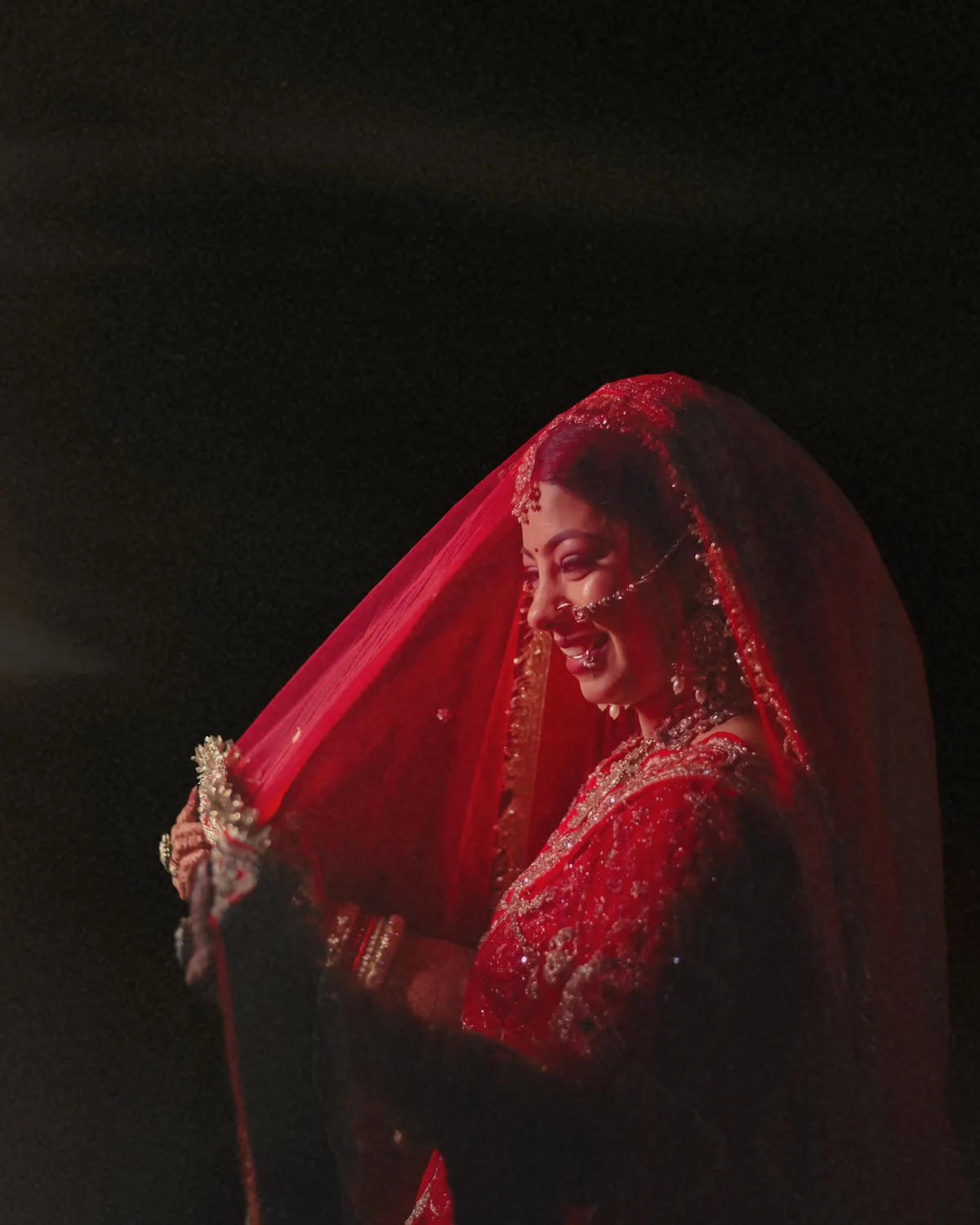 Bride smiling beneath a crimson veil in low light