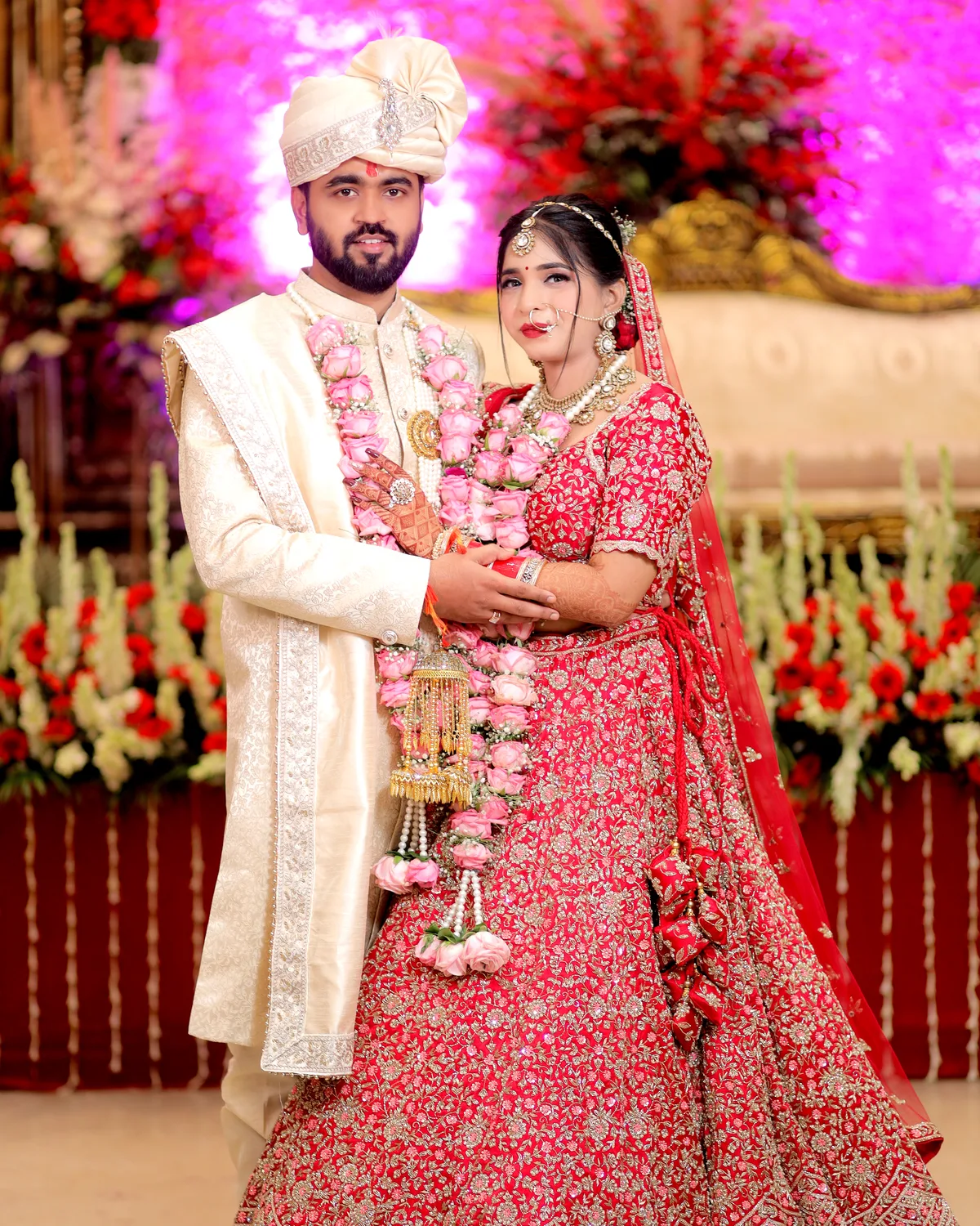 Groom in a white sherwani and bride in a pink lehenga, garlanded, in a flower-decked hall