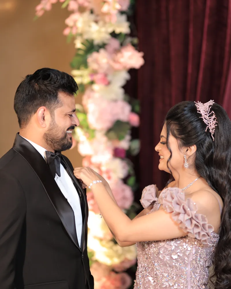 Groom in a tuxedo and bride in a lilac gown by a flower wall
